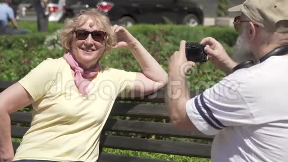 成熟男人在公园的长凳上给他时髦漂亮的妻子拍照视频的预览图