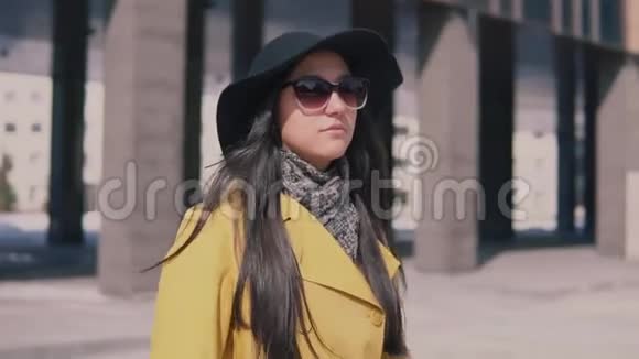 穿着黄色外套戴着深色长发的时髦女孩走在街上摘下眼镜环顾四周视频的预览图