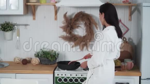 穿着白色浴袍在厨房做饭的年轻女子视频的预览图