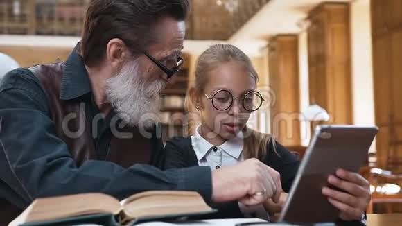 聪明的胡子老爷爷戴着眼镜和他迷人的小孙女坐在图书馆里视频的预览图