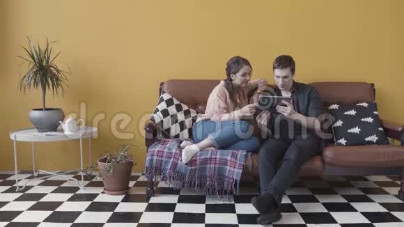 一对夫妇坐在沙发上带着电子平板电脑在一黄色的墙壁和黑白格子地板视频的预览图