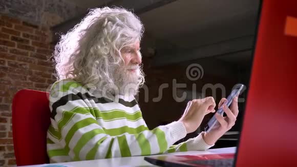 一个可爱的成熟的白种人留着大白胡子和头发坐在砖头旁边的笔记本电脑旁正在刷卡视频的预览图
