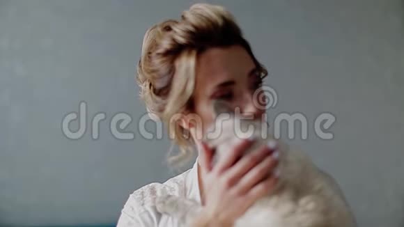 美丽的女孩穿着白色睡衣抱着一只暹罗猫抚摸着他的手他抚摸他的脖子视频的预览图