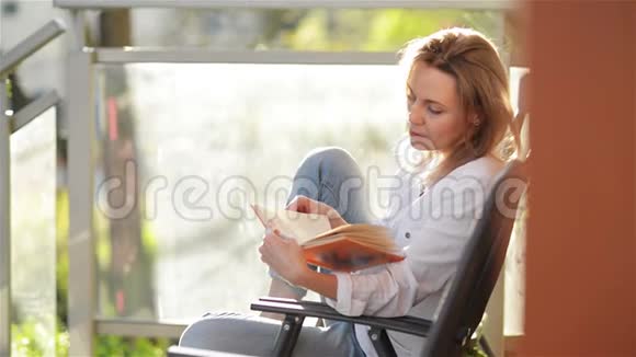 年轻美丽的女孩的肖像与白种人的外观阅读有趣的书坐在阳台上女人在放松视频的预览图