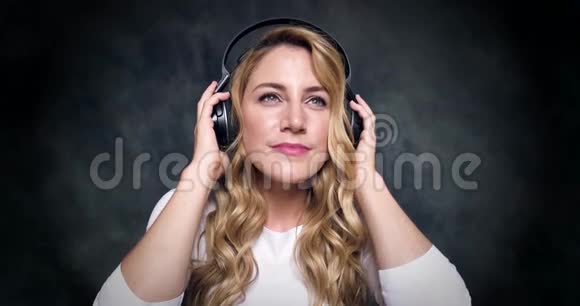 美丽的金发美女在黑暗的背景上听耳机里的音乐视频的预览图
