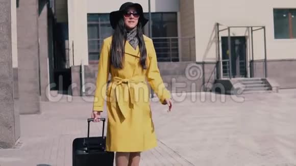 穿着黄色雨衣帽和带着行李的眼镜的时髦女孩赶着出租车视频的预览图