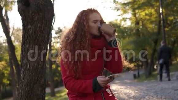 穿着红色外套和围巾的卷发女人站在城市公园里用手机喝咖啡视频的预览图