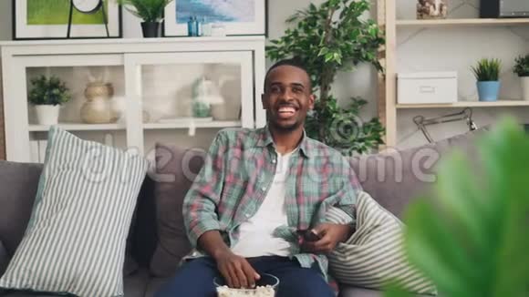 性格开朗的非裔美国学生在电视上看滑稽喜剧在沙发上笑着放松使用遥控器视频的预览图