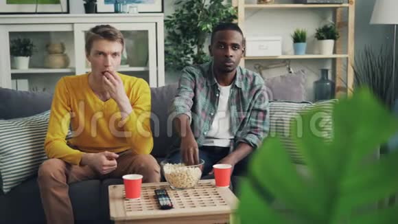 令人不安的年轻人非裔美国人和高加索人的朋友们正在看电视脸上没有感情吃爆米花视频的预览图