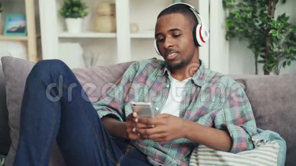 漂亮的非裔美国学生正在使用智能手机触摸屏幕在沙发上用耳机听音乐视频的预览图