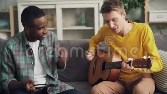 快乐的男人非裔美国人和高加索人的朋友们正在弹吉他用平板电脑和在沙发上放松的笑声视频的预览图