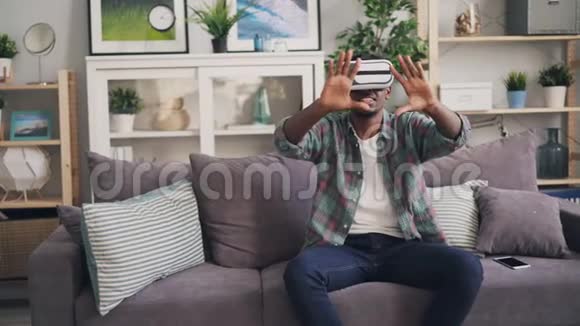 非洲裔美国人使用虚拟现实眼镜笑着坐在沙发上动动手享受着快乐视频的预览图