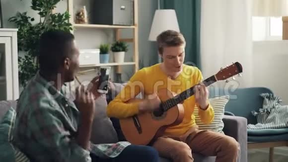 非洲裔美国年轻人拿着现代智能手机拍着他的高加索朋友弹吉他的视频视频的预览图