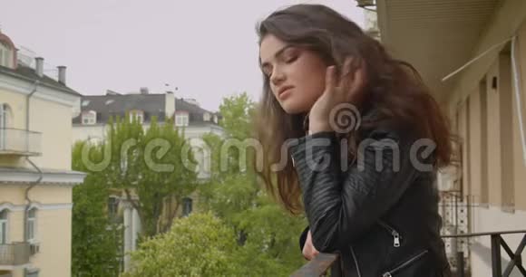 年轻的黑发女孩穿着皮夹克站在阳台上欣赏壮观的绿色城市景色视频的预览图