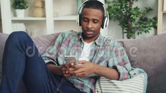 年轻的非裔美国人正在使用智能手机坐在公寓的沙发上通过耳机听音乐视频的预览图