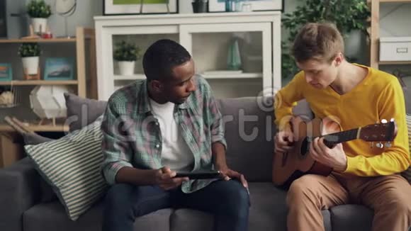 白种人年轻人和他的非裔美国朋友正在一起学习用平板电脑弹吉他视频的预览图