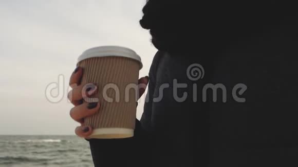 戴着帽子和围巾的女孩从海边的一次性杯子里喝茶视频的预览图