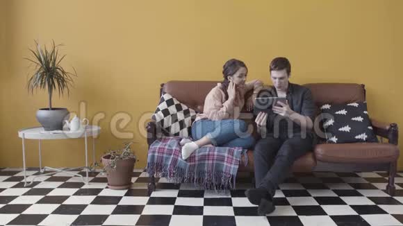 一对夫妇坐在沙发上带着电子平板电脑在一黄色的墙壁和黑白格子地板视频的预览图