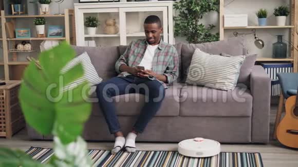 无护理的非裔美国人使用坐在沙发上的机器人吸尘器手里拿着智能手机享受着乐趣视频的预览图