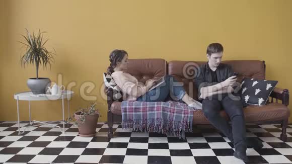 年轻夫妇坐在家里的沙发上沉迷于现代小玩意库存录像使用手机的人视频的预览图