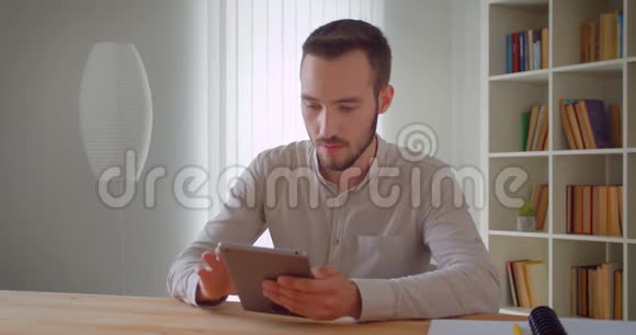 年轻英俊的白种人商人用平板电脑在公寓的室内拍摄的特写肖像上面有书架视频的预览图