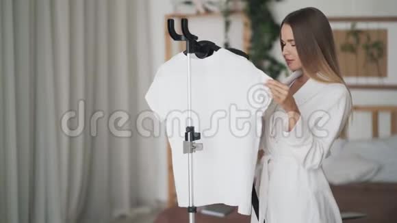 穿着白色浴袍用蒸汽系统熨烫衣服的漂亮年轻女子视频的预览图