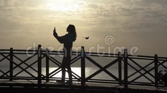 一个年轻女孩在智能手机上拍照日出时站在桥上视频的预览图