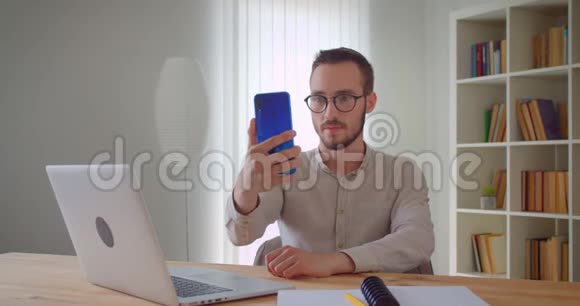 年轻英俊的白种人商务明星坐在笔记本电脑前用手机自拍的特写照片视频的预览图