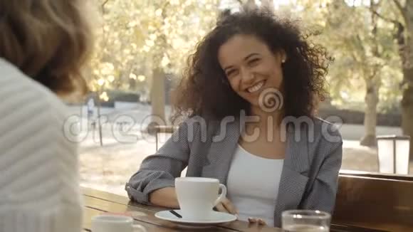 年轻女子在户外餐馆里端着咖啡和朋友聊天视频的预览图