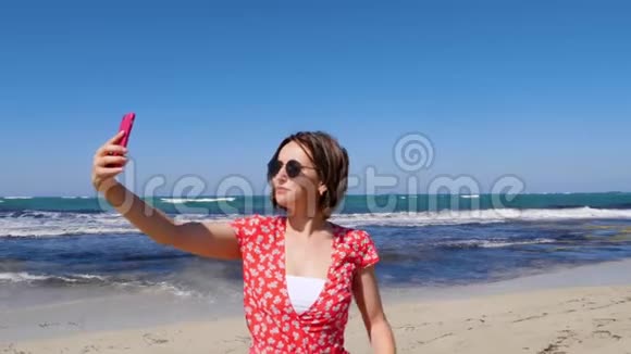 年轻的美女穿着红色连衣裙带着相机手机在海风和海浪的海滩上自拍慢动作视频的预览图
