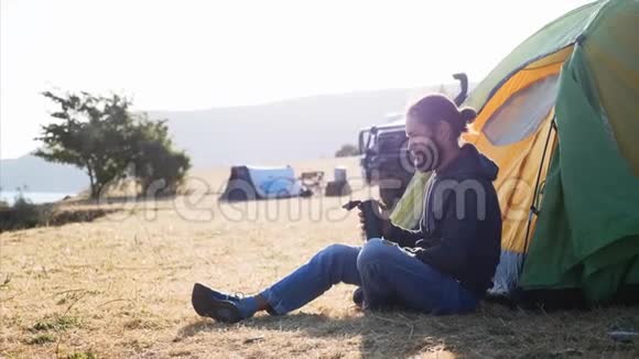 年轻人坐在营地帐篷附近喝着热水瓶里的茶视频的预览图