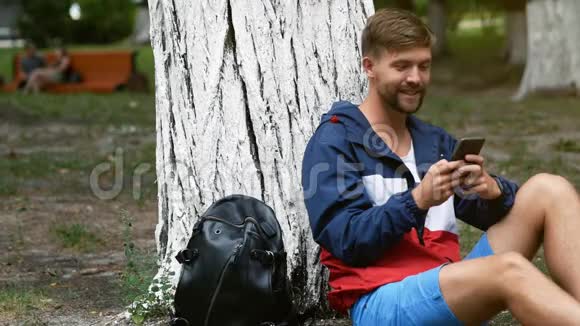 坐在树下的小胡子男人用手机打字学生在公园里聊天视频的预览图