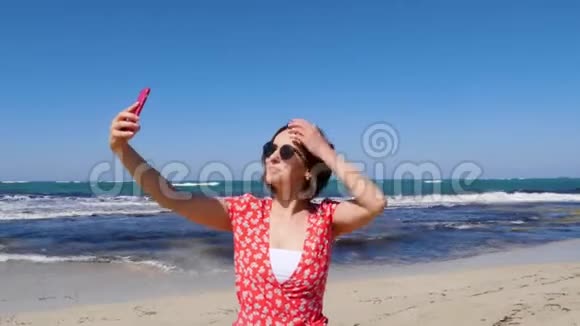 年轻的美女穿着红色连衣裙带着相机手机在海风和海浪的海滩上自拍视频的预览图