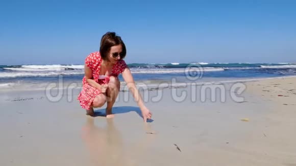 年轻迷人的女人在一个孤独的沙滩上画着心脏标志强烈的波浪撞击着海岸她穿着红裙子晒太阳视频的预览图