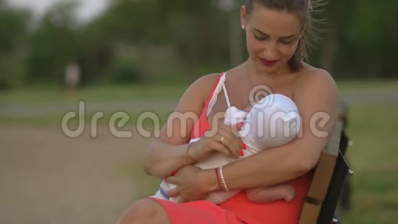母乳喂养年轻的母亲在城市公园里给她的孩子喂奶她坐在长凳上穿着鲜红的连衣裙视频的预览图