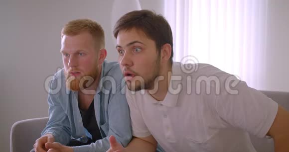 两个英俊的白种人成年男子在电视上看体育节目坐在室内沙发上悲伤的特写镜头视频的预览图