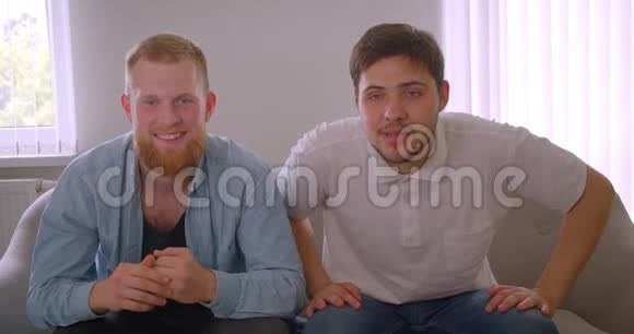 两个英俊的白种人成年男子坐在室内沙发上看足球比赛的特写镜头视频的预览图
