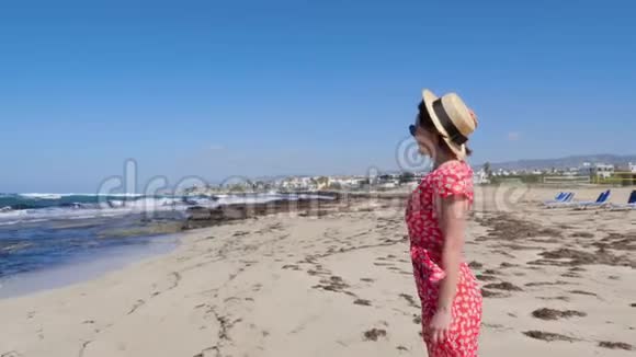 年轻漂亮的女人穿着红色的连衣裙和帽子独自站在空荡荡的海滩上躺着日光浴沙滩有强烈的波浪和风视频的预览图
