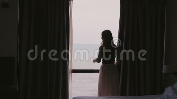 后景年轻幸福的女人用史诗般的海景关上窗帘走回黑暗的酒店公寓慢动作视频的预览图