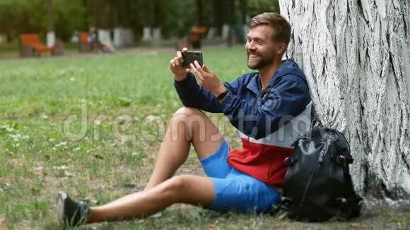 帅哥在公园里坐在树下一边拿着智能手机视频聊天与朋友聊天视频的预览图