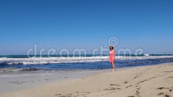 年轻快乐的女人走在海滩上戴着红色的裙子和太阳镜把帽子扔掉强烈的风浪和蓝色的天空视频的预览图