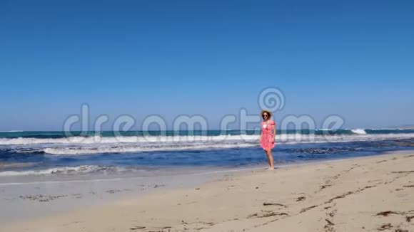 年轻快乐的女人走在海滩上戴着红色的裙子和太阳镜把帽子扔掉强烈的风浪和蓝色的天空视频的预览图