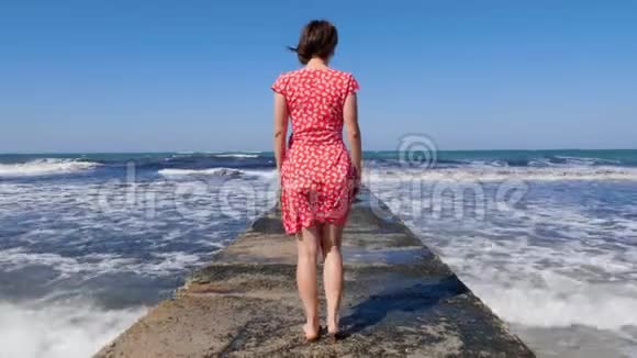 年轻女子赤着脚走在海上码头上而海浪撞击码头后面的镜头视频的预览图