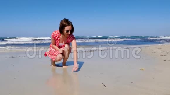 年轻迷人的女人在一个孤独的沙滩上画着心脏标志强烈的波浪撞击着海岸她穿着红裙子晒太阳视频的预览图