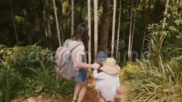 镜头跟随着幸福的年轻妈妈和两个小孩子一起走在陡峭的异国情调的森林小径上慢动作视频的预览图