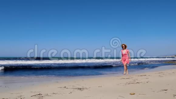 年轻的白种人女子赤脚在沙滩上奔跑把帽子扔在天空中她穿着红色连衣裙和太阳镜视频的预览图