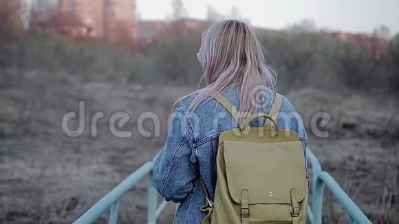 肩上背着背包的女孩在城市荒原上她穿过一座蓝色的小桥从后面看视频的预览图