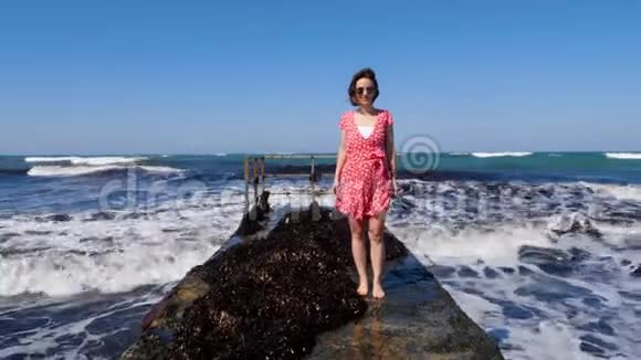 年轻快乐的女人穿着红色的裙子和太阳镜站在码头上海浪拍打着码头视频的预览图
