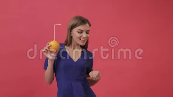 漂亮的女孩穿着裙子喝橙汁用鸡尾酒吸管跳舞紫色背景工作室视频的预览图