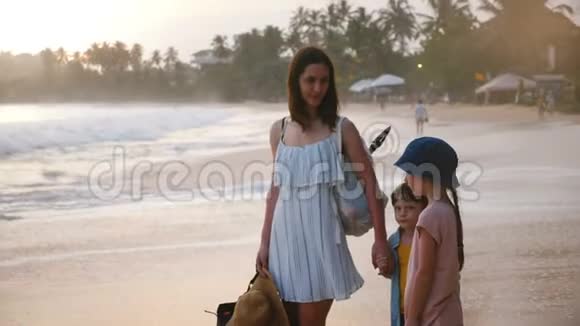 快乐微笑的年轻妈妈和两个小孩子在美丽的夕阳下在异国情调的热带海滩上交谈视频的预览图